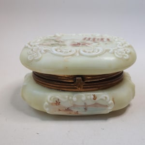 May include: An oval-shaped, off-white milk glass trinket box featuring a hinged brass metal closure. The lid and base are decorated with embossed scrollwork patterns and hand-painted landscape scenes in muted tones of brown, green, and blue.