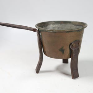 May include: A small copper pot with a metallic interior, featuring three dark iron legs riveted to the sides and a long, slender iron handle extending from the rim. The exterior surface shows a weathered, brownish-orange patina with dark spots.