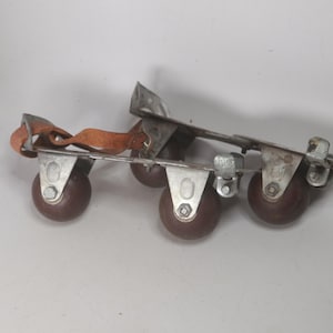 May include: Vintage roller skates with brown wheels and metal frames. The skates have a brown leather strap for securing the foot. The wheels are a dark brown color, and the metal shows signs of wear.