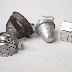 May include: A collection of vintage tea infusers in various shapes and designs. Includes a teapot-shaped infuser with a chain, a mesh strainer, a cone-shaped infuser, and a house-shaped infuser with a hinged lid. All are made of metal.