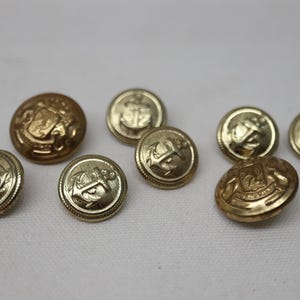 May include: A collection of seven round gold-toned metal buttons. Five of the buttons feature a raised anchor design, while two larger buttons display a detailed crest with an anchor motif. Each button has a textured border and a metallic finish.