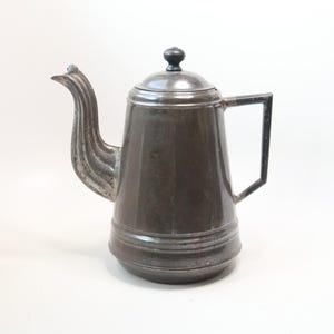 May include: A vintage metal coffee pot with a dark, aged finish. The pot features a curved spout, a lid with a small knob, and a rectangular handle. The design suggests a classic, antique style, suitable for kitchen decor.