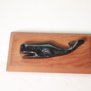 May include: A black whale sculpture is mounted on a rectangular, wooden plaque. The whale is detailed with an open mouth and a curved tail. The wood has a reddish-brown hue, and the sculpture is set against a white background.