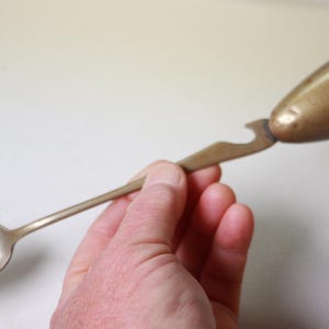 May include: A vintage brass spoon with a bottle opener and a conical end. The spoon has a long handle and a rounded bowl. The bottle opener is located near the conical end. The brass has a warm, aged patina.