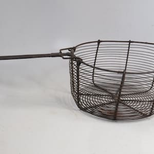 May include: A vintage, rusty metal wire basket with a long handle. The basket is oval-shaped and constructed of closely spaced, curved wires. The handle is attached to the side of the basket. The basket is likely used for frying or straining food.