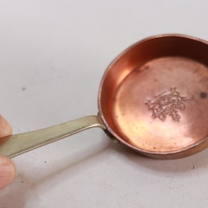 May include: A small copper saucepan with a brass handle. The interior of the pan has a decorative embossed design. The pan is held by a hand.
