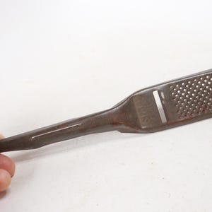 May include: A vintage, rusty metal grater with a long handle and a perforated grating surface. The grater has a small hole at the end for hanging. The grater is held by a hand.