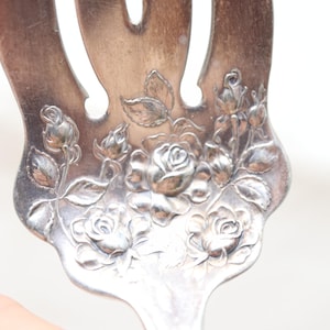 May include: A silver-toned metal serving fork featuring an embossed floral pattern of roses and leaves on the handle. The metal surface shows signs of natural oxidation and patina, with detailed engravings of blooming rose buds and open flowers.