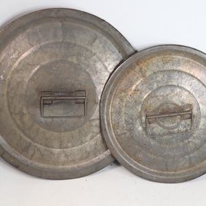 May include: Two circular, galvanized metal lids of different sizes, each featuring a raised concentric ring design and a central rectangular metal handle. The surfaces exhibit a weathered, mottled grey finish with signs of oxidation and wear.