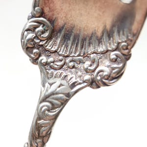 May include: A silver-toned metal spoon handle featuring an intricate embossed floral and scrollwork pattern. The surface displays a tarnished patina with dark oxidation in the recessed areas, highlighting the detailed relief of the metalwork.