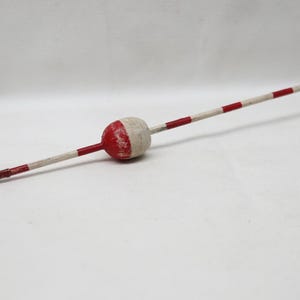 May include: A vintage fishing float featuring a central oval-shaped wooden body painted in red and white. The float has a long, slender stem extending from both ends, decorated with alternating red and white bands, and a small metal wire loop at one end.