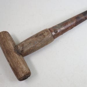 May include: A T-shaped tool featuring a weathered wooden handle and a long, cylindrical metal shaft. The metal component shows signs of oxidation and discoloration, while the wooden handle displays a natural, textured grain and worn finish.