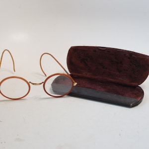May include: Antique round eyeglasses with wire frames and clear lenses. The glasses are next to an open, burgundy-colored glasses case. The case has a velvet interior and a dark exterior.
