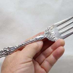 May include: A silver-toned metal fork featuring an intricate embossed floral pattern along the handle and the base of the tines. The four-tined utensil has a polished finish with detailed scrollwork and leaf motifs decorating the entire length.
