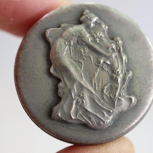 May include: A round, metallic button with a matte gray finish. The center features a raised, detailed relief sculpture depicting a figure in flowing garments reaching toward a smaller figure amidst stylized foliage and organic shapes.