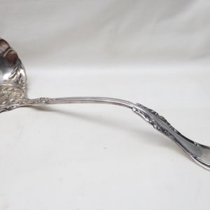 May include: A silver-toned metal soup ladle featuring a deep, rounded bowl and a long, curved handle. The handle and the base of the bowl are decorated with intricate, embossed floral and scrollwork patterns throughout the entire surface.