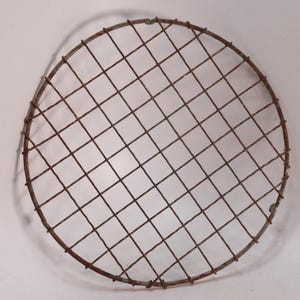 May include: A circular, rusty metal grill grate with a grid pattern. The grate is made of intersecting wires, forming a series of small squares. The metal has a weathered appearance, suggesting age or outdoor use. The overall shape is round.