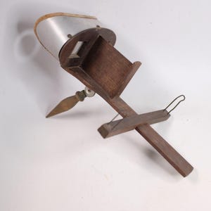 May include: A vintage wooden stereoscope viewer featuring a dark brown wooden frame, a sliding card holder with a thin metal wire support, and a curved metal hood with a wooden top edge. The device includes a small wooden handle for holding.