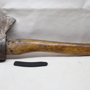 May include: A weathered steel hatchet with a curved wooden handle and a dark, worn metal head. The axe head features a visible stamp on the side. A small, curved black rubber or plastic piece lies horizontally beneath the handle.