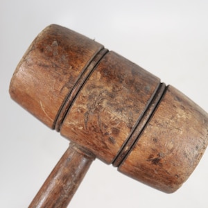 May include: A brown wooden mallet featuring a cylindrical head with two distinct horizontal grooves encircling the center. The wood displays a weathered, textured surface with natural grain patterns and signs of wear throughout the handle and head.
