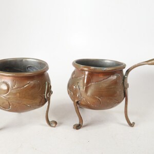May include: Two copper vessels with a metallic finish, each featuring three curved legs and embossed leaf-like patterns on the exterior. One vessel is a round bowl, while the other is a small pot with a single extended handle attached to the side.