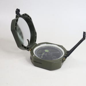 May include: An olive green military-style sighting compass with a hinged lid featuring an integrated mirror. The device includes a circular bubble level, a rotating bezel with white markings, and a black folding sighting arm on the side.
