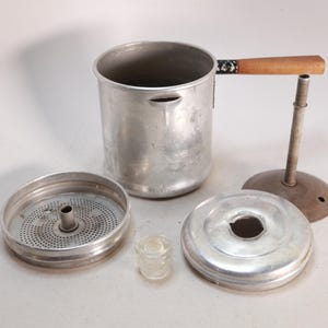 May include: An aluminum coffee percolator with a wooden handle and various components. The set includes a pot, filter basket, lid, and a glass insert. The percolator is designed for brewing coffee and has a vintage aesthetic.