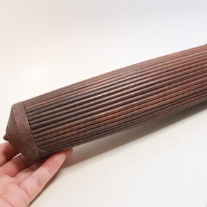 May include: A cylindrical wooden rolling pin featuring a deeply ribbed, fluted texture along its entire length. The object has tapered, pointed ends with a dark brown finish and a consistent, repeating vertical groove pattern across the surface.