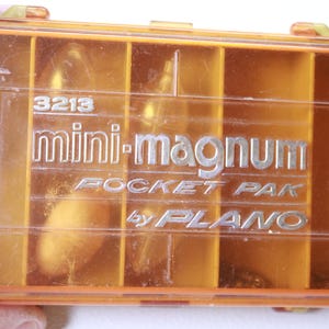 May include: An orange plastic Mini-Magnum Pocket Pak by Plano. The clear lid reveals several compartments. The text "3213" is above the "mini-magnum" logo. The words "POCKET PAK" and "by PLANO" are also visible.