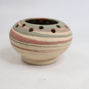 May include: A round ceramic flower frog with a squat, bulbous shape and a flat base. The exterior features horizontal marbled bands in muted shades of sage green, dusty rose, and cream. The top surface has a series of circular holes for stems.