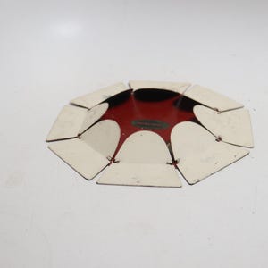 May include: A circular metal object featuring a deep red center and a series of white, petal-like flaps arranged around the perimeter. The flaps are connected to the central red base, creating a flower-like structure with a flat, low-profile design.