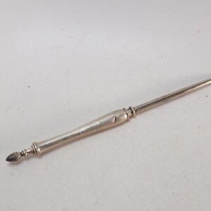 May include: A silver-toned metal button hook featuring a slender, tapered handle with a decorative, textured finial at the end. The tool has a smooth, polished finish and a curved hook at the opposite end for fastening buttons on clothing items.