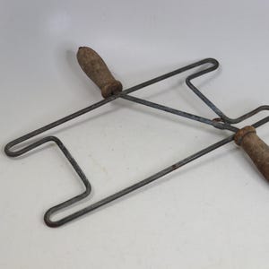 May include: A vintage kitchen tool featuring a rectangular metal wire frame with two wooden handles. The metal frame is dark grey with signs of oxidation, and the two wooden handles are brown, textured, and show visible signs of wear.