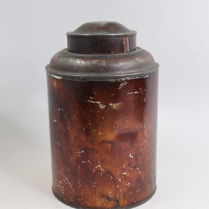 May include: A vintage, cylindrical metal container with a lid. The container is primarily brown with areas of wear and discoloration, suggesting age. The lid is a darker shade of brown. The overall aesthetic is rustic and antique.