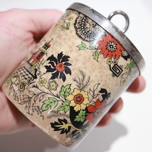 May include: A vintage, cylindrical container with a silver rim and handle. The container is decorated with a floral pattern in red, yellow, and black on a beige background. The design includes a pagoda-like structure.