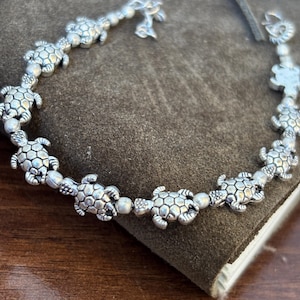 Könnte beinhalten: Ein silberfarbenes Armband mit einer Reihe von aneinandergereihten Meeresschildkröten-Anhängern. Jeder Schildkrötenanhänger zeigt ein detailliertes Panzer-Muster mit strukturierter Oberfläche und kleinen runden silbernen Perlen.