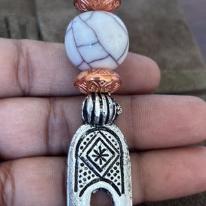 Puede incluir: Una horquilla para el cabello con una base de metal plateado con un patrón de diamante grabado y un motivo de estrella central. La parte superior está decorada con dos cuentas texturizadas de color cobre que flanquean una cuenta blanca grande y redonda con vetas de color marrón oscuro.