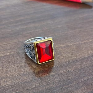 May include: A silver-toned ring featuring a large, rectangular, faceted red stone set in a gold-toned bezel. The band displays an intricate Greek key pattern along the sides with a textured, stippled finish on the metal surface.