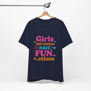 May include: Navy blue t-shirt with the text "Girls just wanna HAVE FUN.. .ctions" in pink, orange, and teal. The text is surrounded by stars and other graphics. The t-shirt is hanging on a wooden hanger.