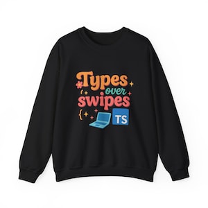 Peut inclure: Sweat-shirt noir avec l'inscription "Types over Swipes" en orange, rose et blanc. Le motif comprend un ordinateur portable et les lettres "TS" dans un carré bleu. Le sweat-shirt est de style col rond.