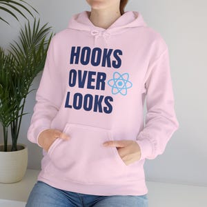 May include: A light pink hoodie with a front pocket and a drawstring hood. The front of the hoodie features the text "HOOKS OVER LOOKS" in dark blue, with a light blue atom symbol. The person is wearing blue jeans.