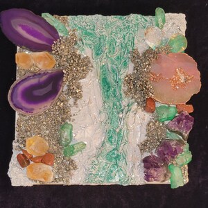May include: A mixed media art piece featuring a variety of crystals and stones, including amethyst, agate, and pyrite. The piece is set on a white background with a green and white textured surface.