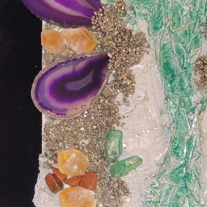 May include: A close-up of a mixed media art piece featuring purple agate slices, clear quartz crystals, and green glass shards on a white background with gold glitter.