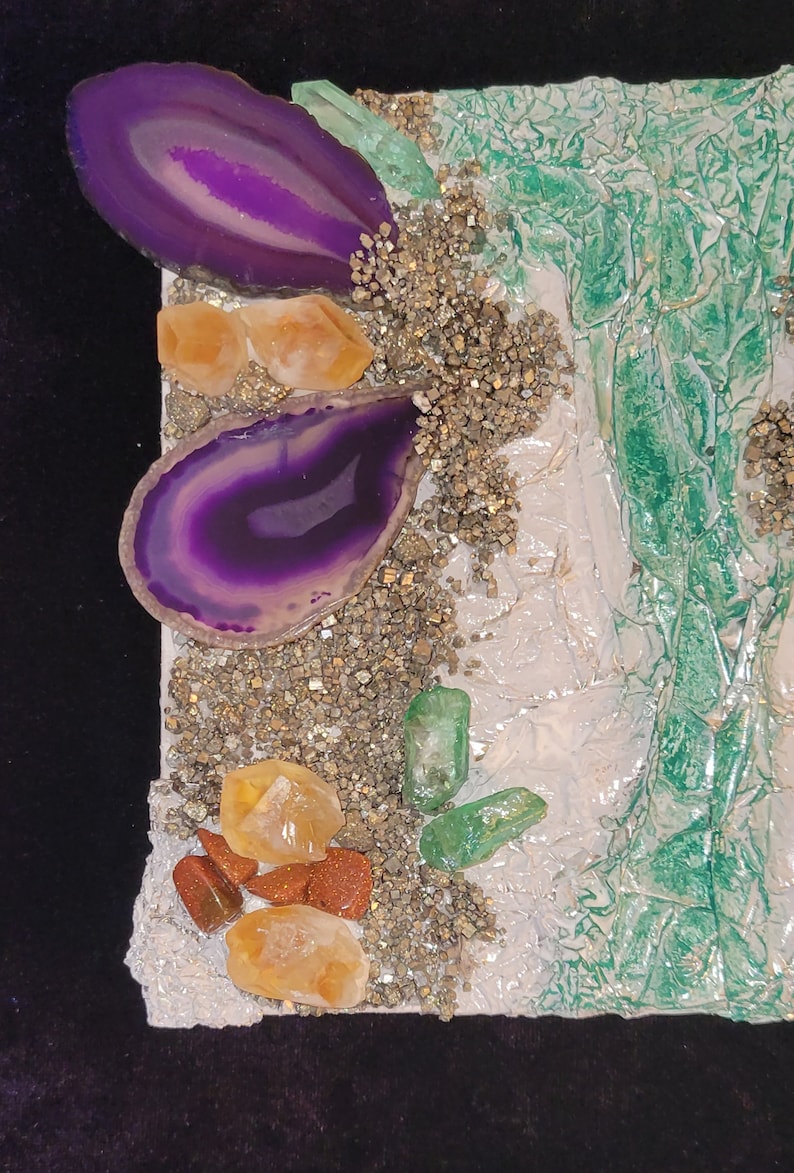 May include: A close-up of a mixed media art piece featuring purple agate slices, clear quartz crystals, and green glass shards on a white background with gold glitter.