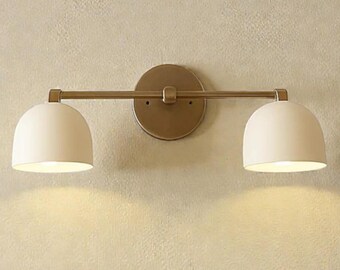 May include: A wall-mounted light fixture with two off-white, dome-shaped shades. The fixture has a brass-colored arm and a circular backplate. The lights are positioned above a bed with white pillows and a tan pillow.