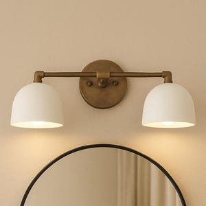 May include: A bathroom vanity set featuring a round black-framed mirror and a two-light fixture. The light fixture has a brass finish and two white dome-shaped shades. The mirror is centered below the lights.