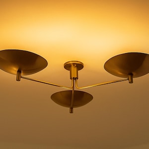 May include: A brass-colored ceiling light fixture with three disc-shaped shades. The fixture has a central stem and three arms extending to hold the shades. The light is warm and diffused.