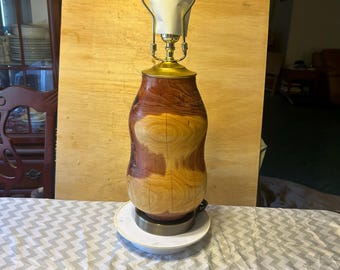 May include: A wooden table lamp with a unique, organic shape. The lamp base is made of polished wood with a mix of light and dark brown tones. The lamp has a brass-colored neck and a white shade with the number "4" written on it.