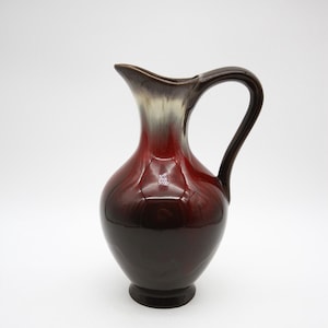 May include: A glossy, vintage ceramic jug with a dark brown handle and base, transitioning to a deep red body and a light brown and white rim. The jug has a rounded body and a curved spout.