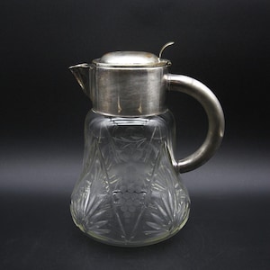 May include: A clear glass pitcher with a silver metal lid and handle. The pitcher has a decorative etched design of leaves and grapes. The handle is a curved, semi-circular shape. The pitcher is on a black background.